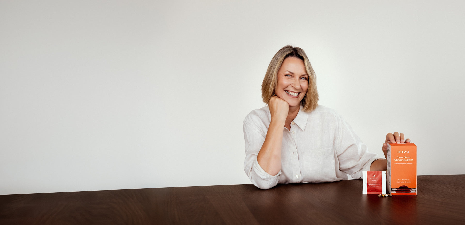 A smiling woman in a white blouse sits at a dark wooden table, holding an orange Nuwa Supplement box and standing next to a red daily supplement pack and 4 capsules. The background is a clean, solid off-white wall.