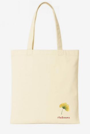 A cream-colored canvas tote bag with two shoulder straps, featuring a small yellow ginkgo leaf graphic and the text '@hellonuwa' printed near the bottom right corner, set against a plain white background.
